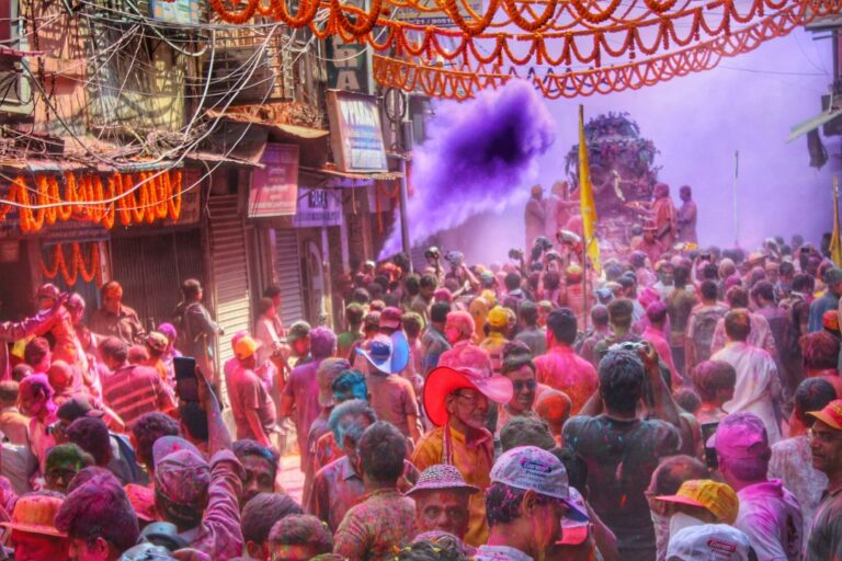 Lively street scene of Holi festivities with people dancing, splashing water, and celebrating with colors under the spring sunlight.